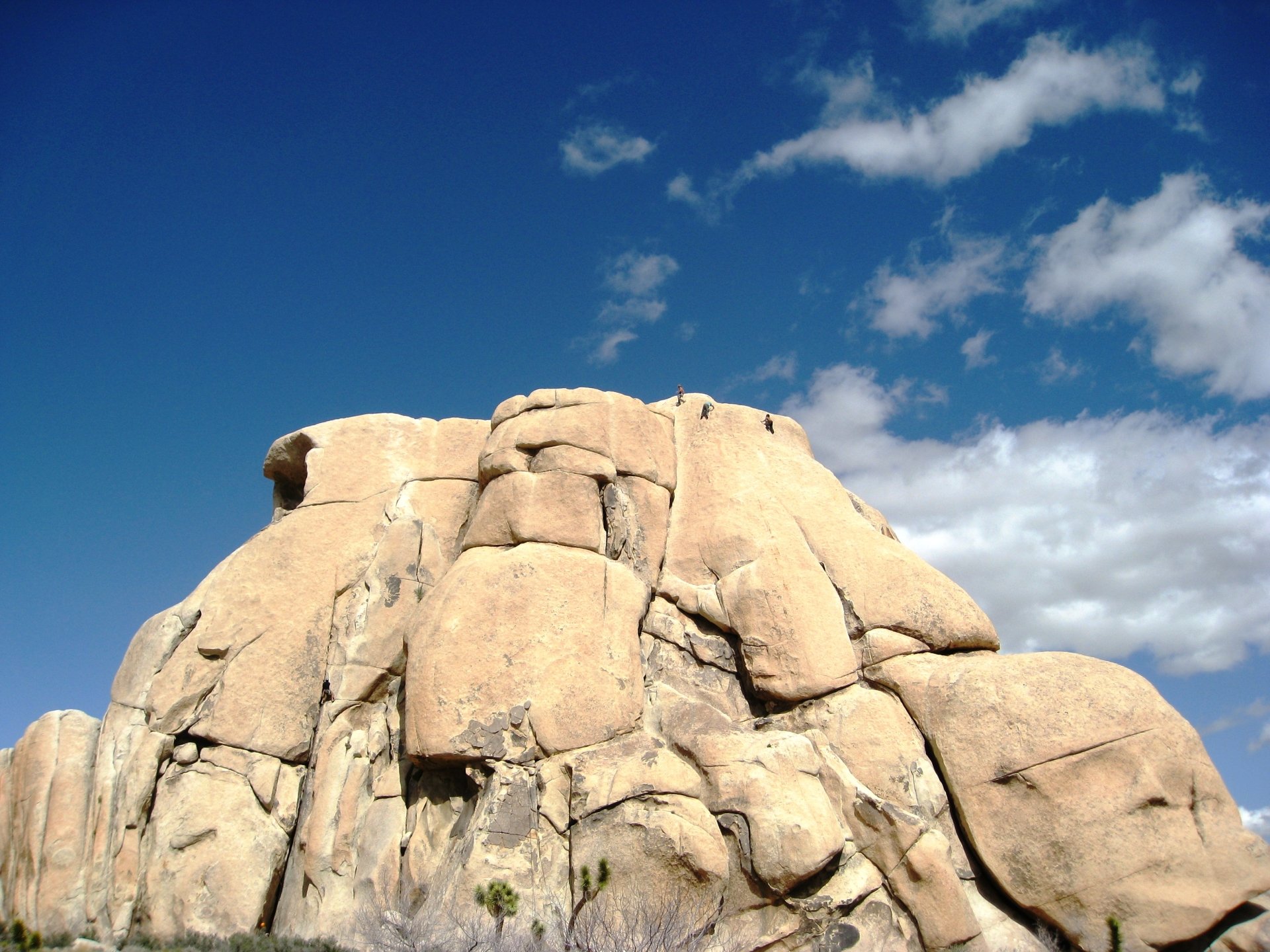 Massive rock formation rises from a desert landscape under a clear blue sky with a few scattered clouds.