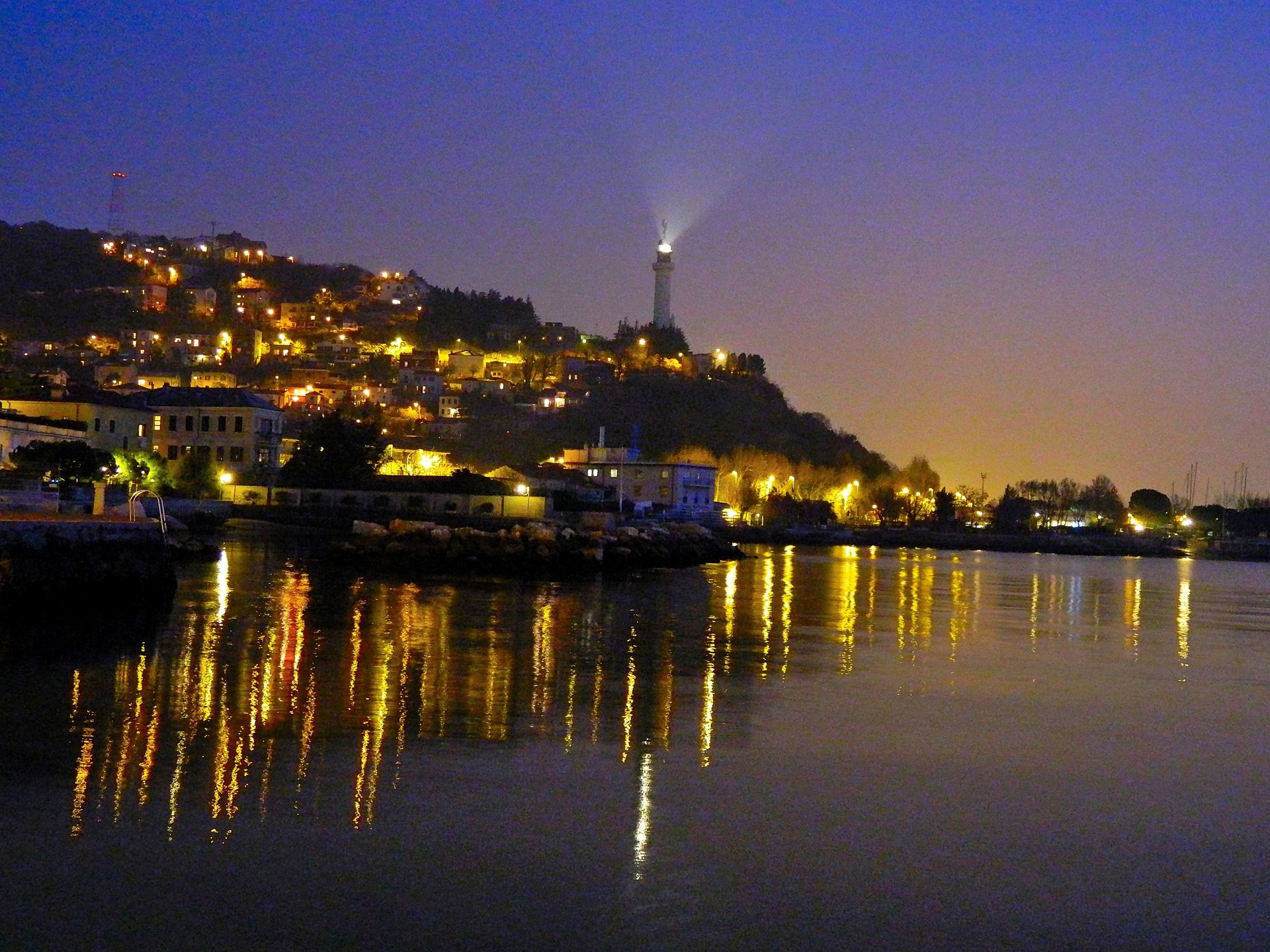 A man-made cityscape illuminated at night, with lights reflecting on the calm water and a statue or monument visible on a hill in the background.