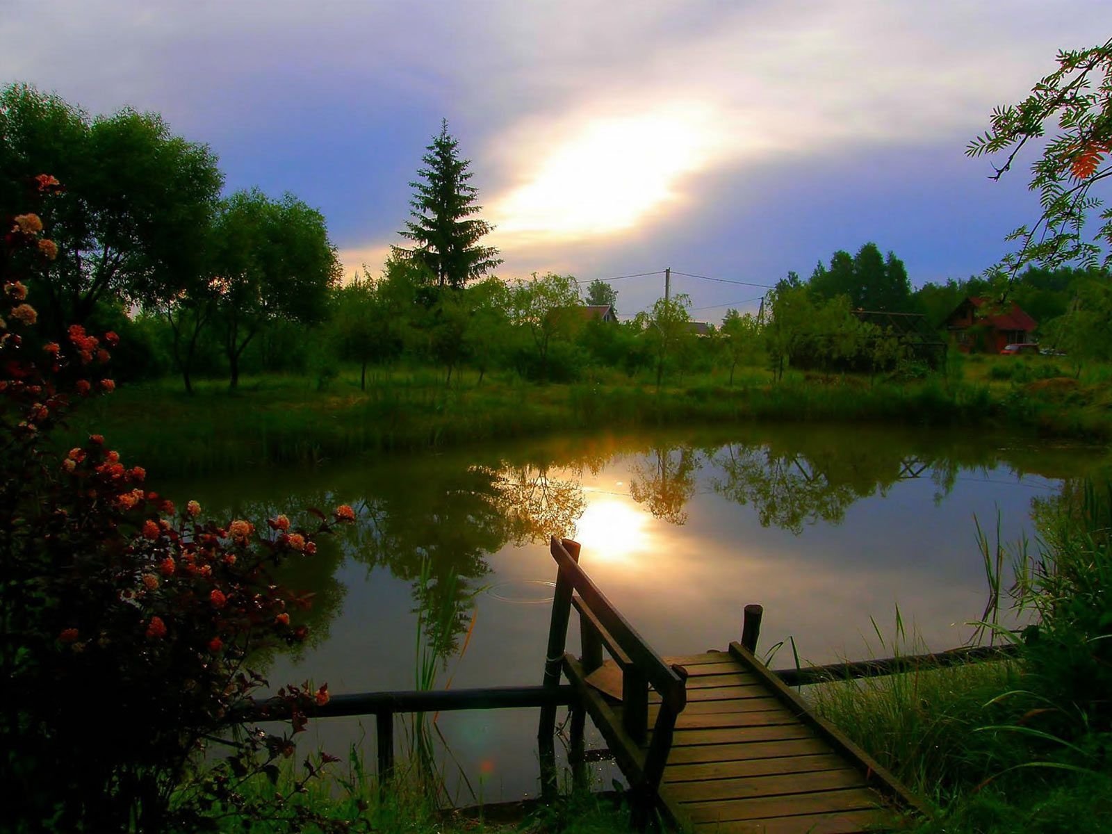 A serene lake reflects the sky at sunset, bordered by lush greenery and flowers, with a wooden dock leading out over the calm water.