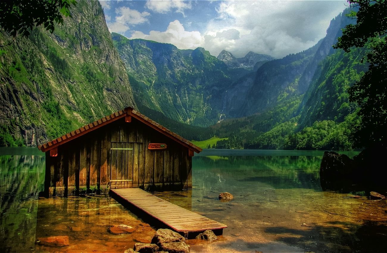 A serene lake scene featuring a wooden cabin on the water, surrounded by towering mountains and lush greenery under a cloudy sky. The calm reflection adds to the tranquil atmosphere.