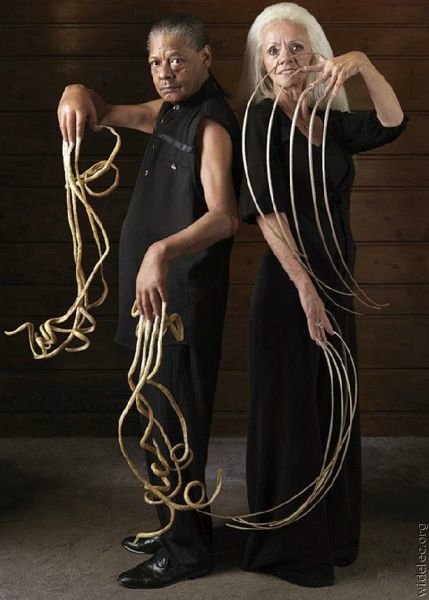 A striking photograph of two people showcasing their exceptionally long nails, standing back-to-back against a wooden backdrop, highlighting their unique and artistic presentation.