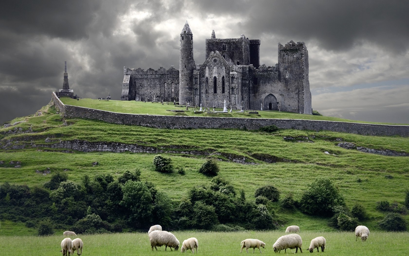 A ruined man-made church stands on a hill under a cloudy sky, with sheep grazing peacefully on the green grass below.