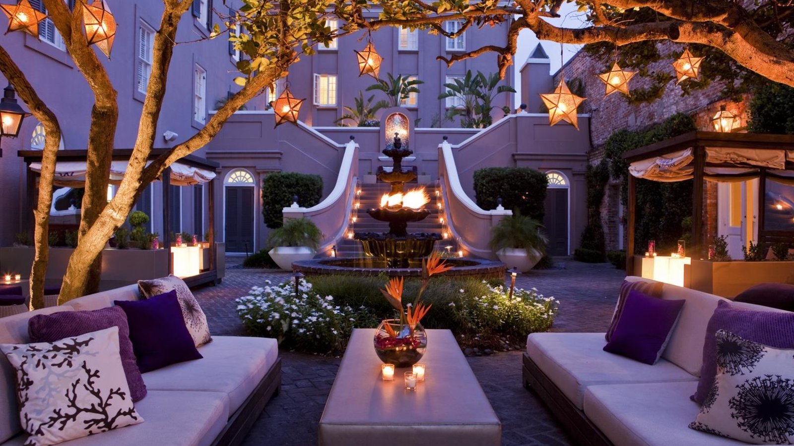 A cozy outdoor space featuring elegant seating, a central fountain, and decorative lights among lush greenery, creating a serene, man-made retreat.