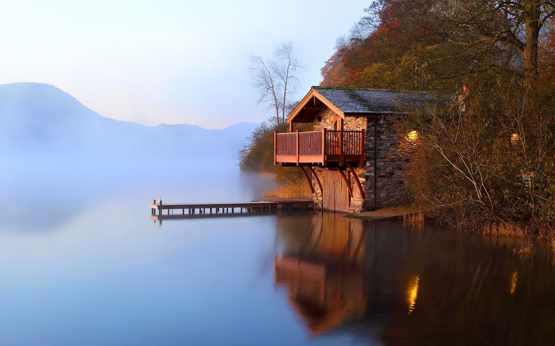 A serene boathouse sits on the calm waters of a lake, surrounded by misty hills and trees, reflecting the tranquil scene in the water at dawn.