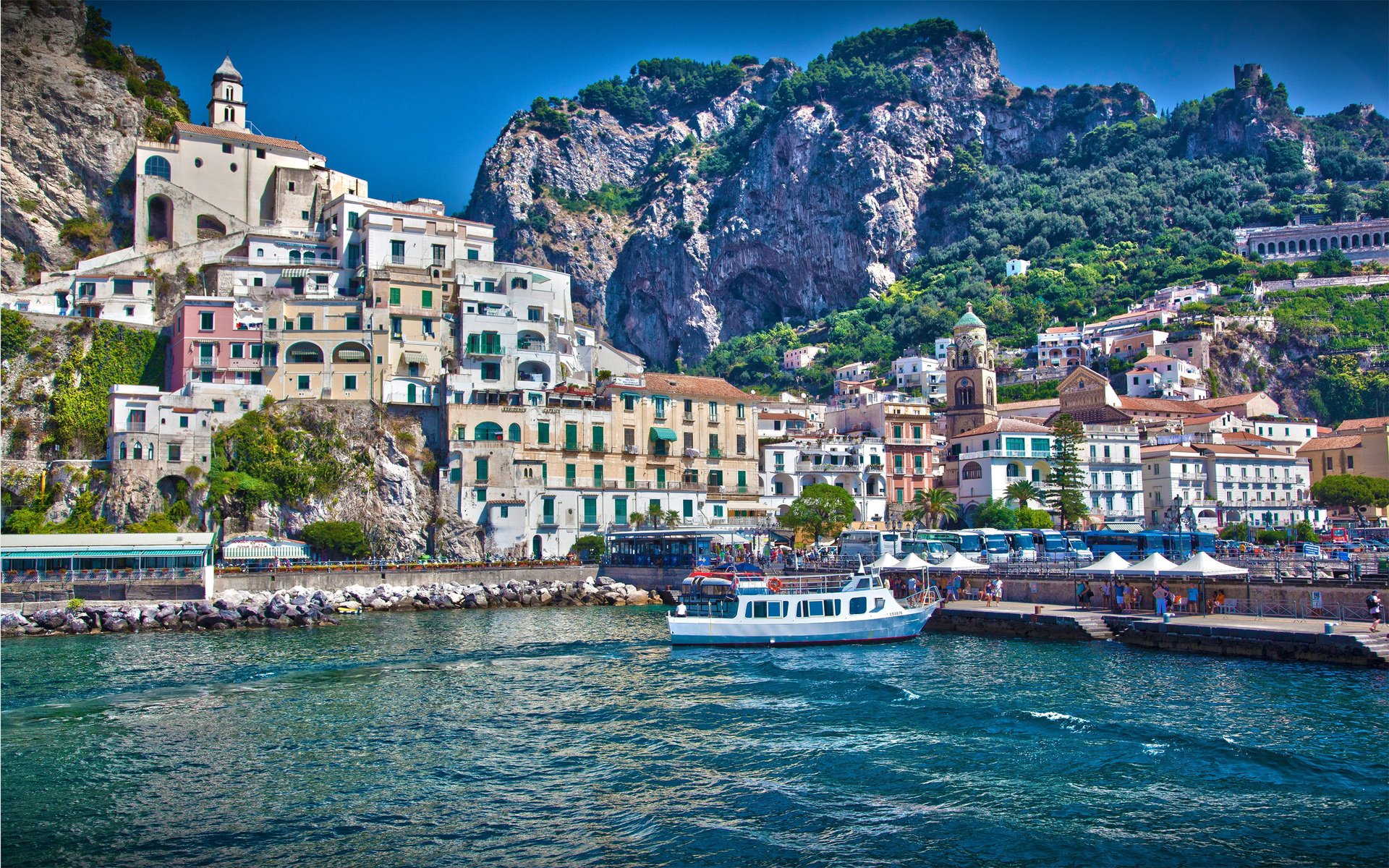 A scenic man-made village built along a coastal hillside with colorful buildings, a church, and boats docked by the waterfront under a clear blue sky.
