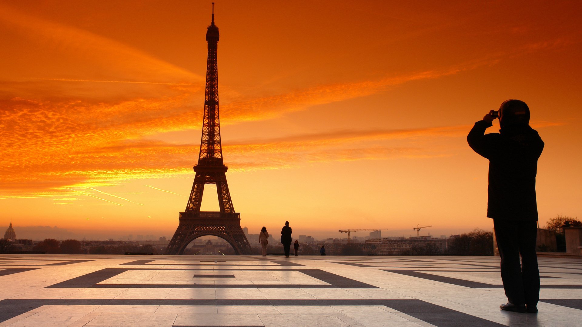 A silhouette of a man photographing the Eiffel Tower at sunset, with vibrant orange skies and two figures walking in the foreground, showcases the iconic man-made landmark.