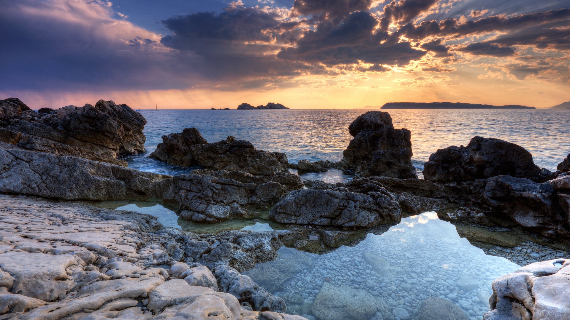 Nature seascape at sunset: rocky shoreline with tidal pools reflecting a colorful sky and distant islands on a calm sea.