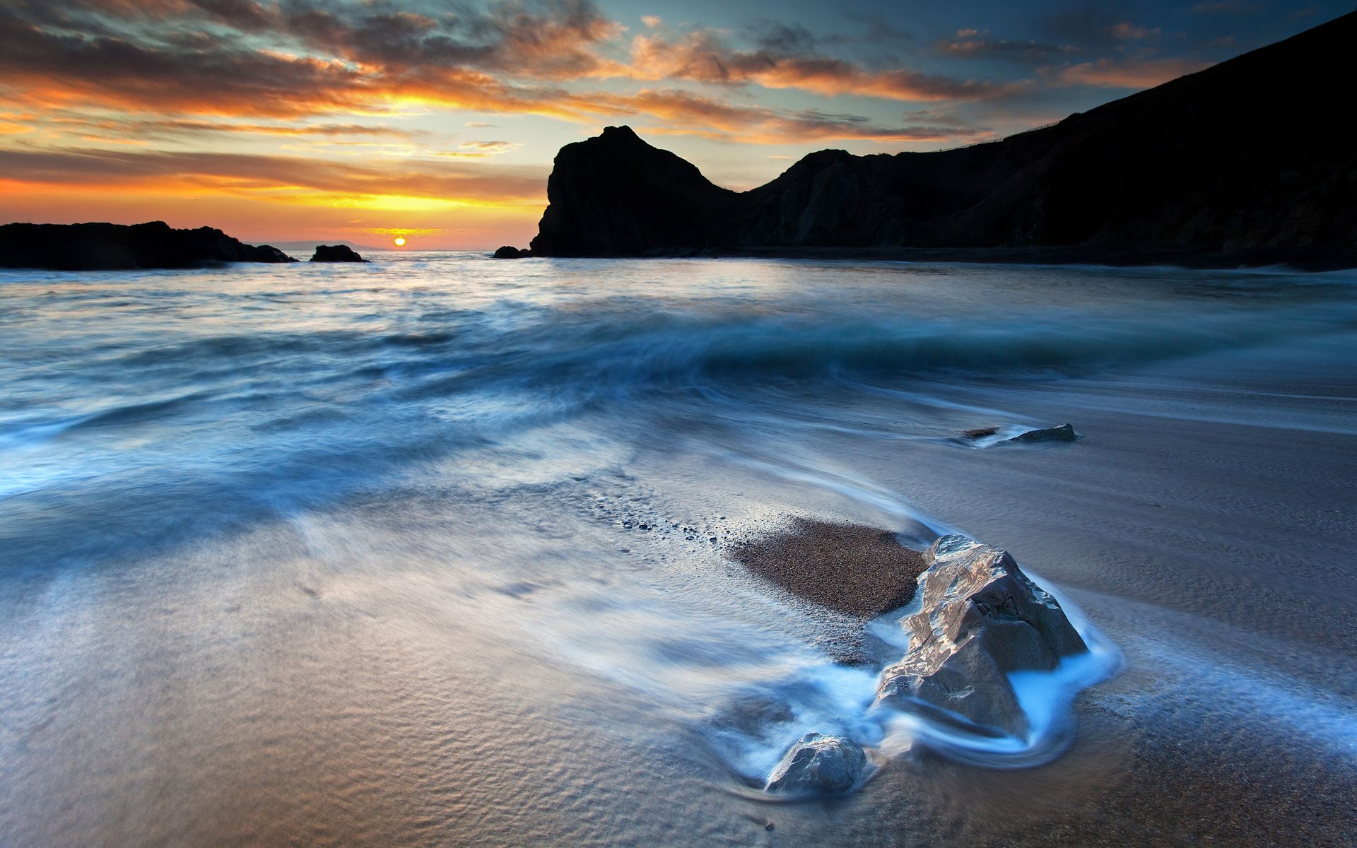 A stunning coastal scene at sunset, showcasing gentle waves lapping against sandy shores, with rocky formations in the background, capturing the beauty of nature.