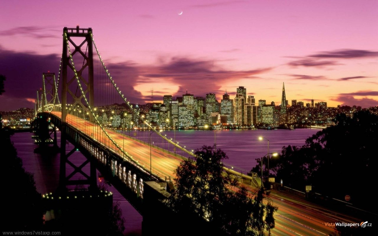 Golden Gate Bridge illuminated at sunset, spanning over water with the city skyline glowing under a vibrant purple and pink sky.