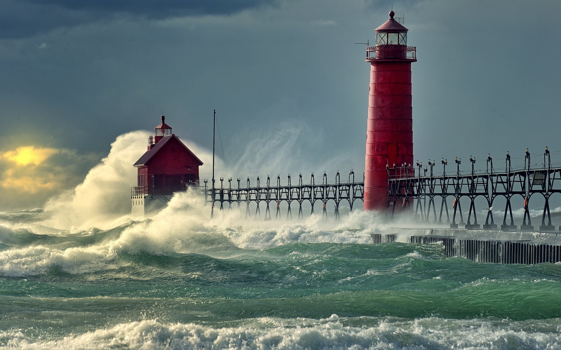 Red man-made lighthouse and keeper's house on a long pier as towering waves crash, spraying green sea under a stormy sky.