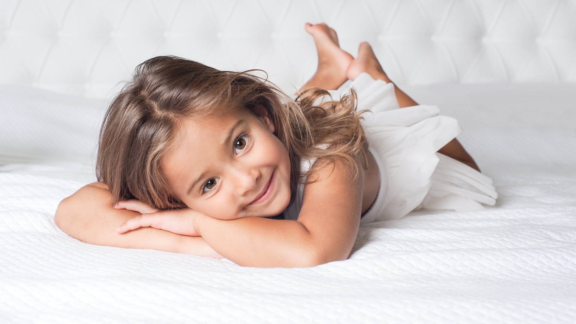 A smiling child lies on a white bedspread, resting her chin on her arms. She has long hair and wears a light outfit, exuding a playful and joyful spirit.