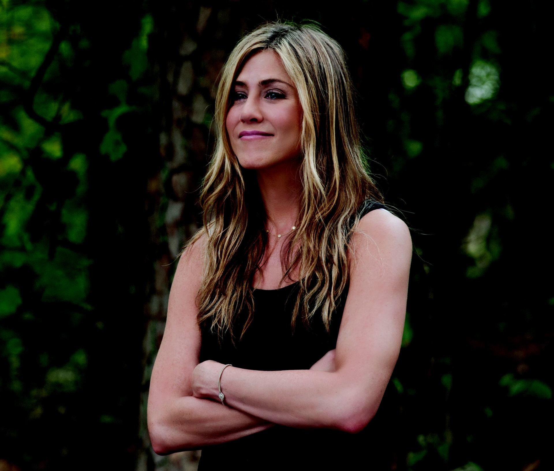 A smiling woman with long, wavy hair stands outdoors with crossed arms, capturing a moment from the movie Wanderlust. The natural setting adds to the relaxed vibe.