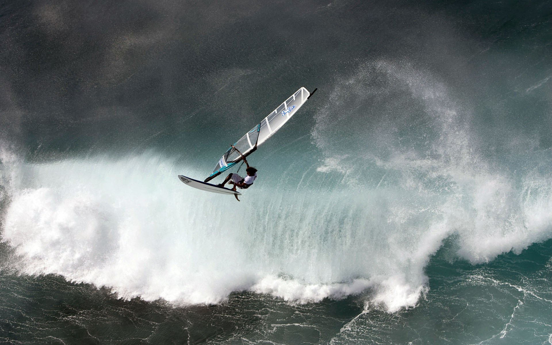 Windsurfing: athlete airborne on a sailboard above a crashing ocean wave in a dynamic sports action shot.