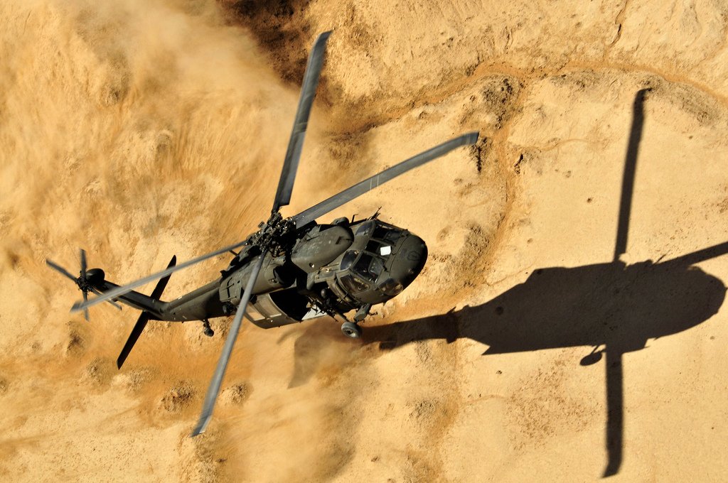 A military Sikorsky UH-60 Black Hawk helicopter flies over rocky terrain, casting a distinct shadow below.