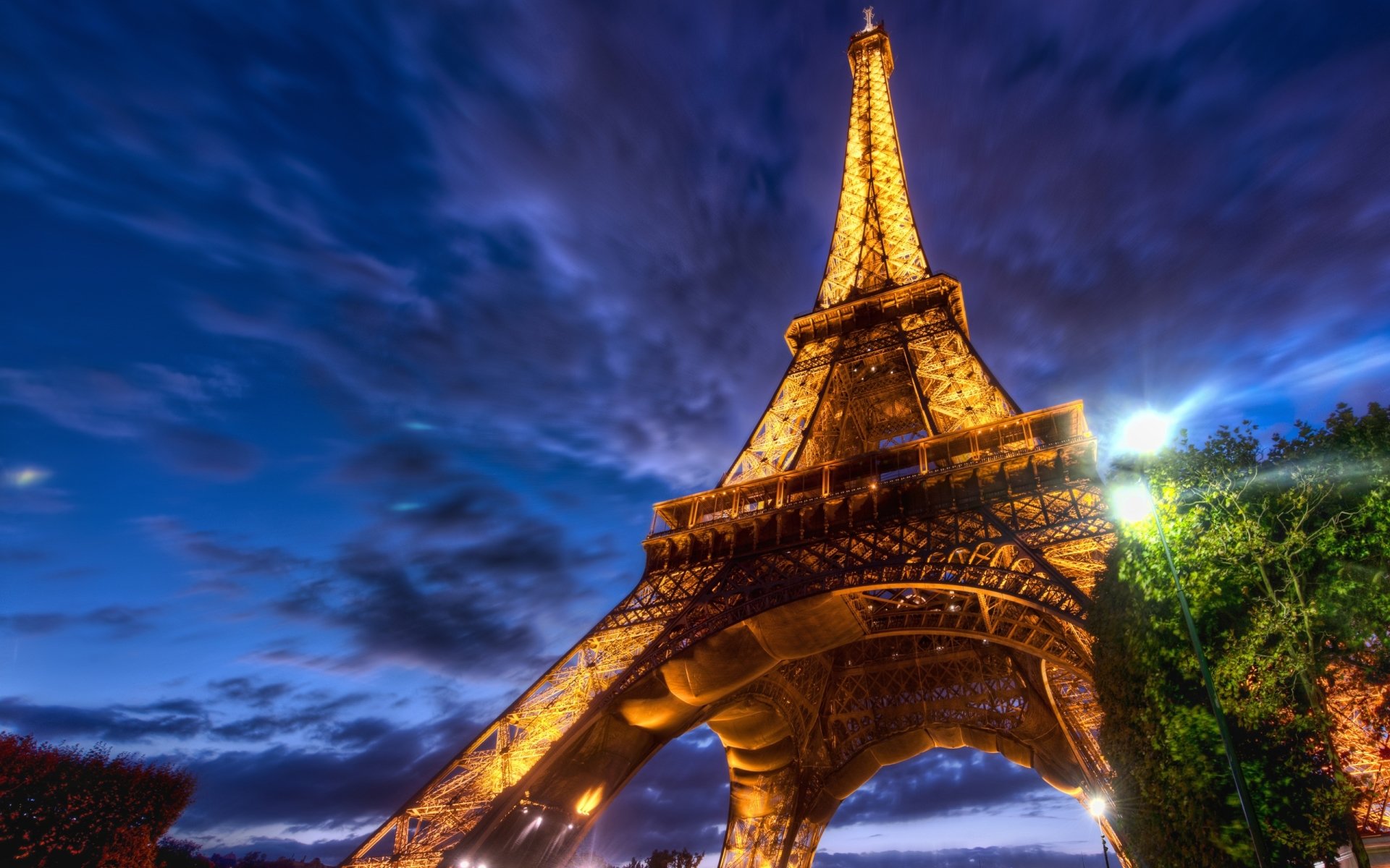 The Eiffel Tower illuminated at dusk, showcasing its intricate ironwork and towering presence against a dramatic sky filled with clouds.