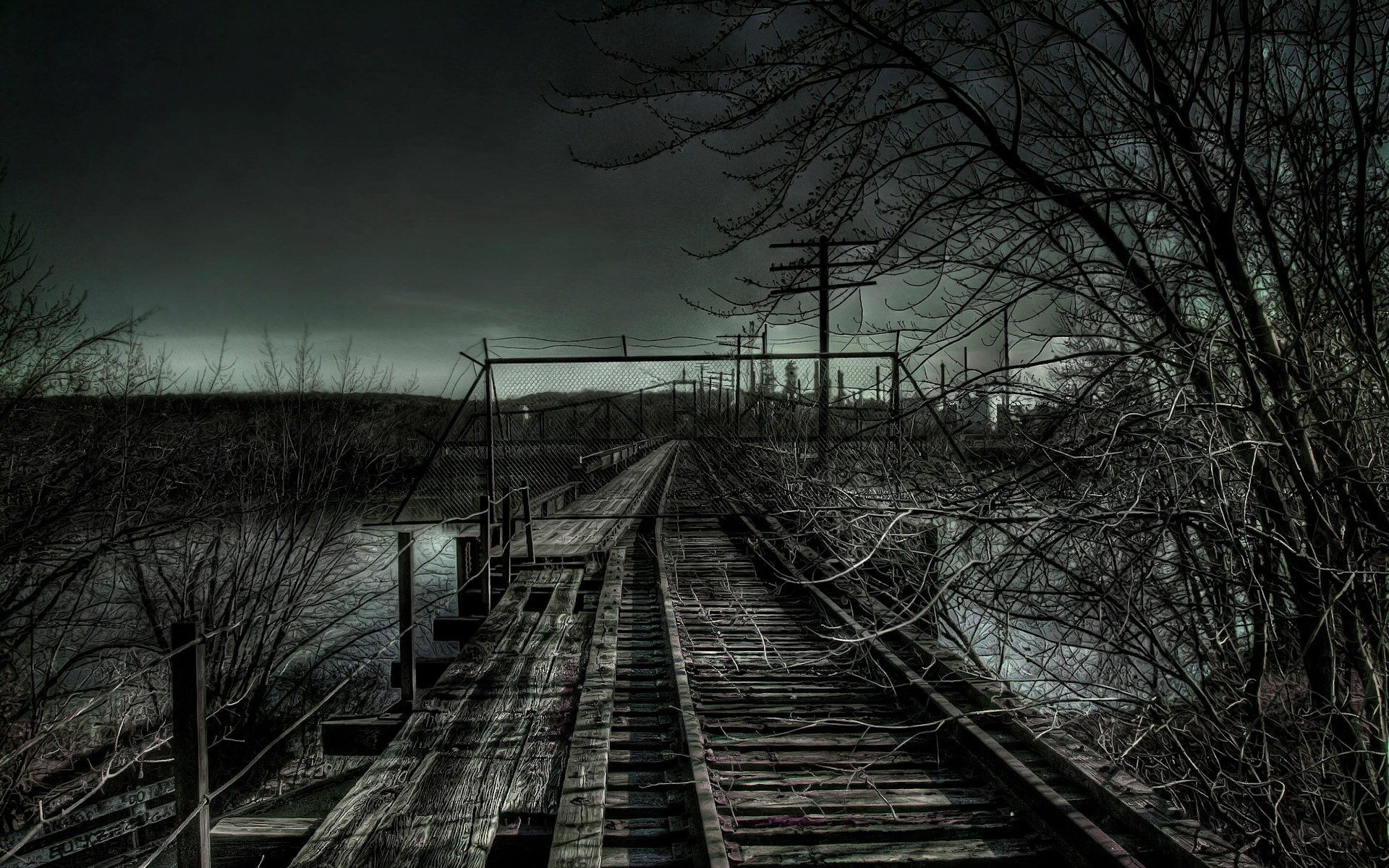 A dimly lit railroad track stretches into the distance, flanked by bare trees and a cloudy sky, showcasing a hauntingly tranquil man-made landscape.