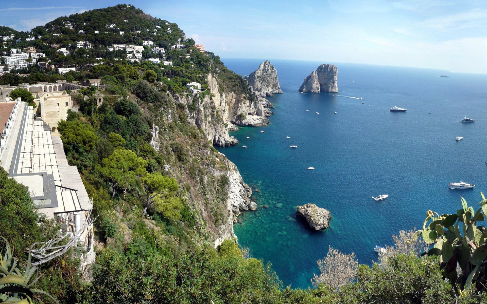 Cliffside nature seascape of turquoise sea with rocky stacks, boats, and white houses dotting verdant hills under a clear blue sky.