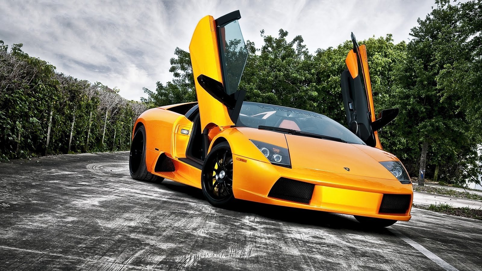A striking orange Lamborghini Murcielago parked on a winding road, showcasing its signature scissor doors against a backdrop of lush greenery and a dramatic sky.