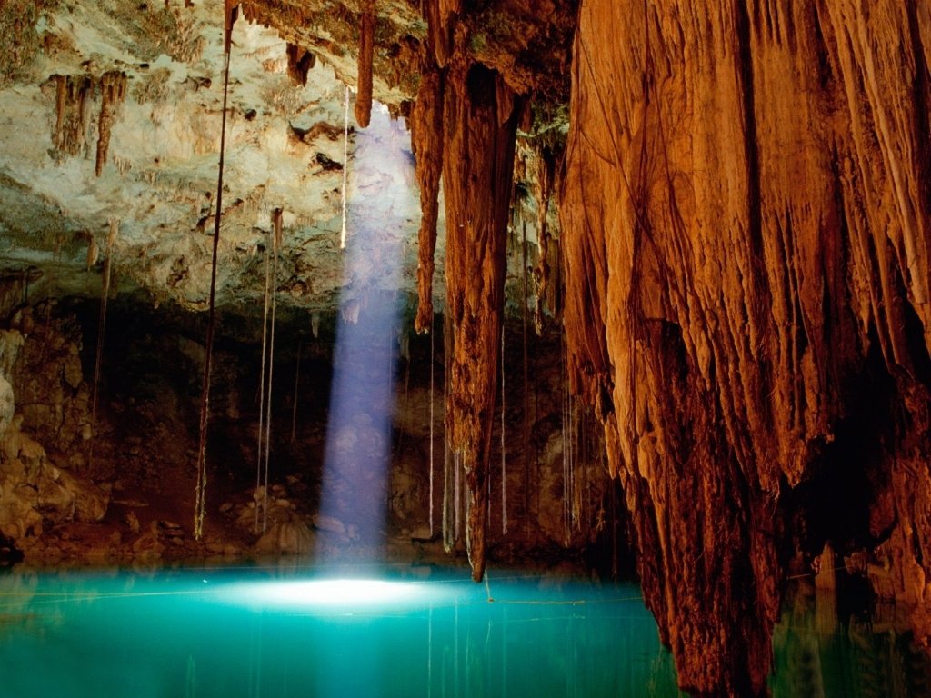 Sunlight streams through a natural opening into a cave, illuminating turquoise water surrounded by stalactites and stalagmites in this striking nature scene.
