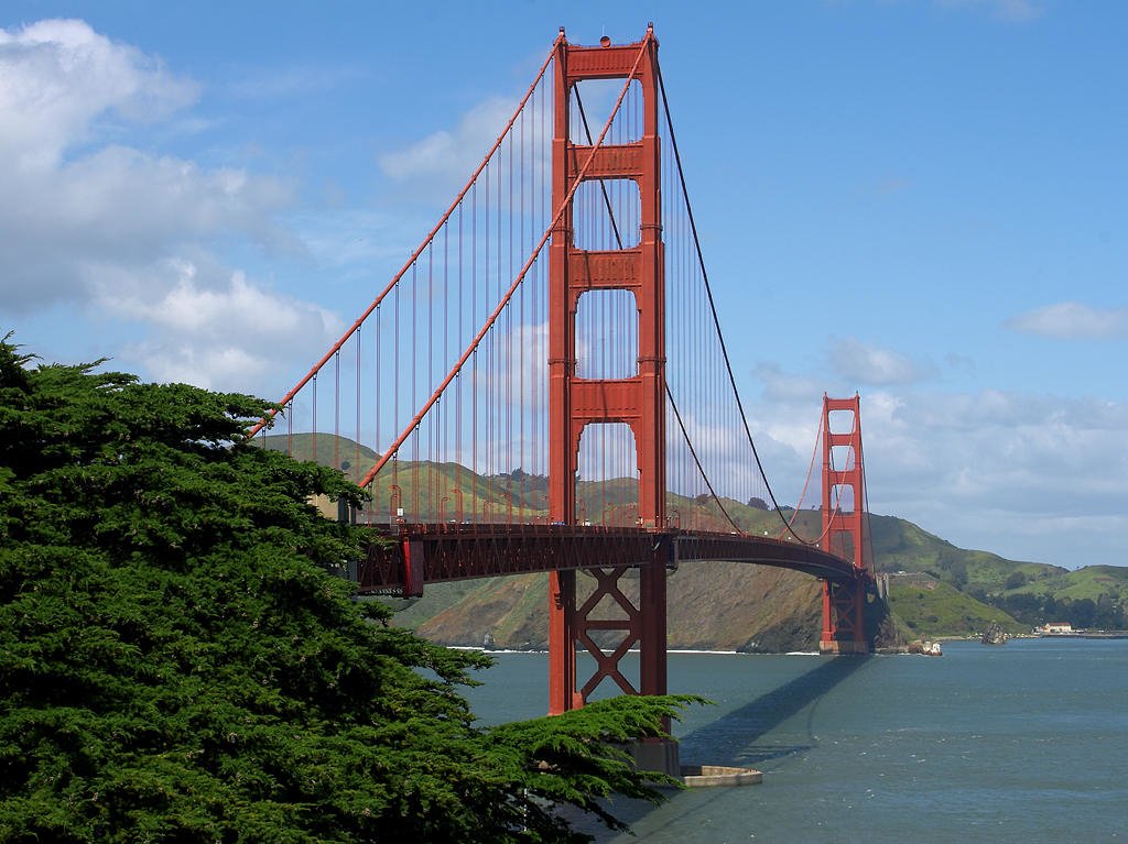 View of the iconic man-made Golden Gate Bridge stretching across the water with hills and greenery in the background under a partly cloudy sky.