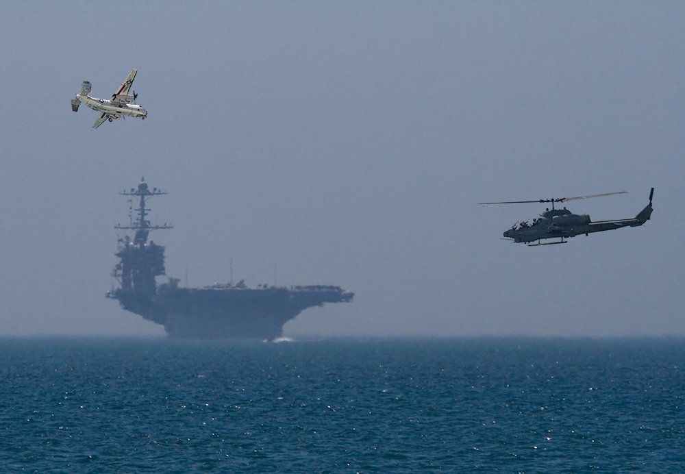 A military aircraft carrier looms in the background, while a helicopter and an aircraft maneuver above the ocean, showcasing naval aviation in action.