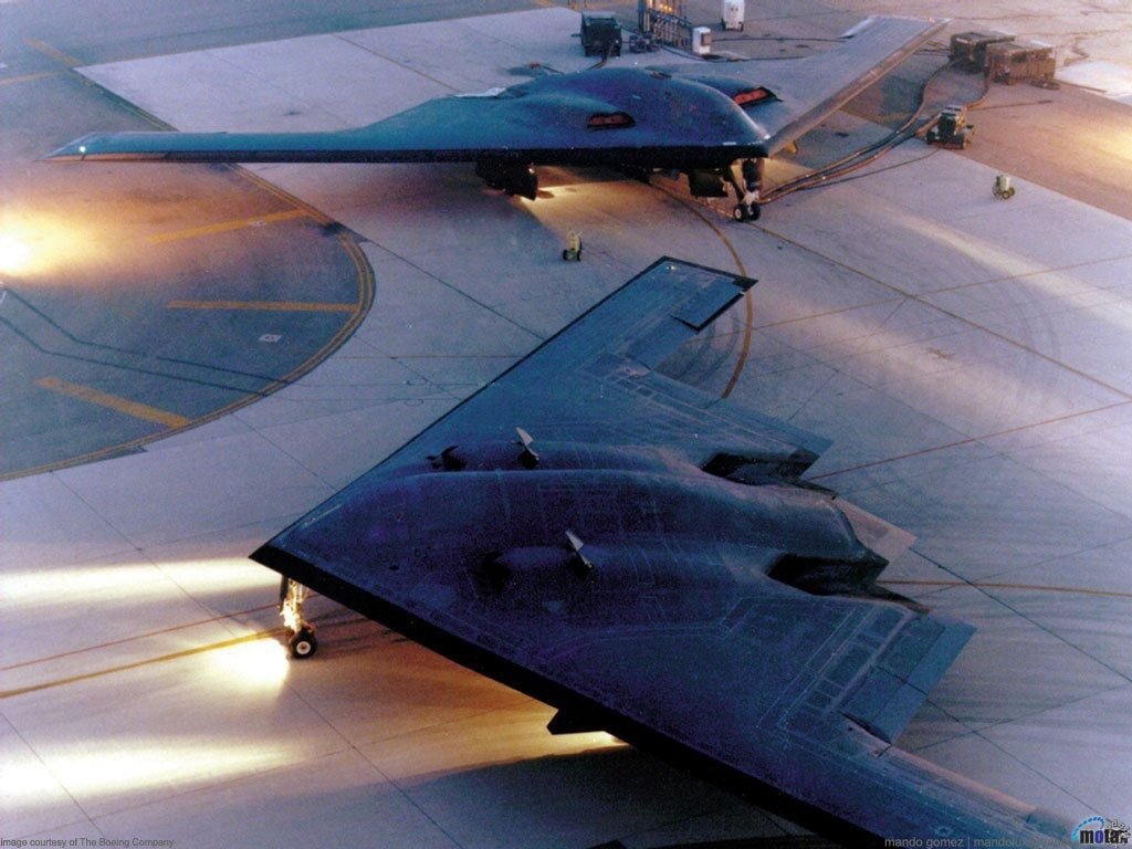 Two military aircraft with stealth designs are parked on a tarmac during dusk, illuminated by ground lights.