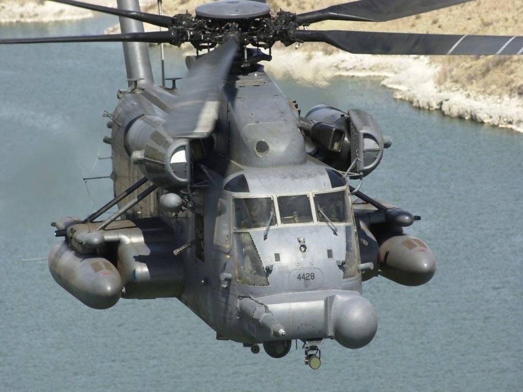 A military helicopter in flight over a body of water, showcasing its robust design and mounted equipment.