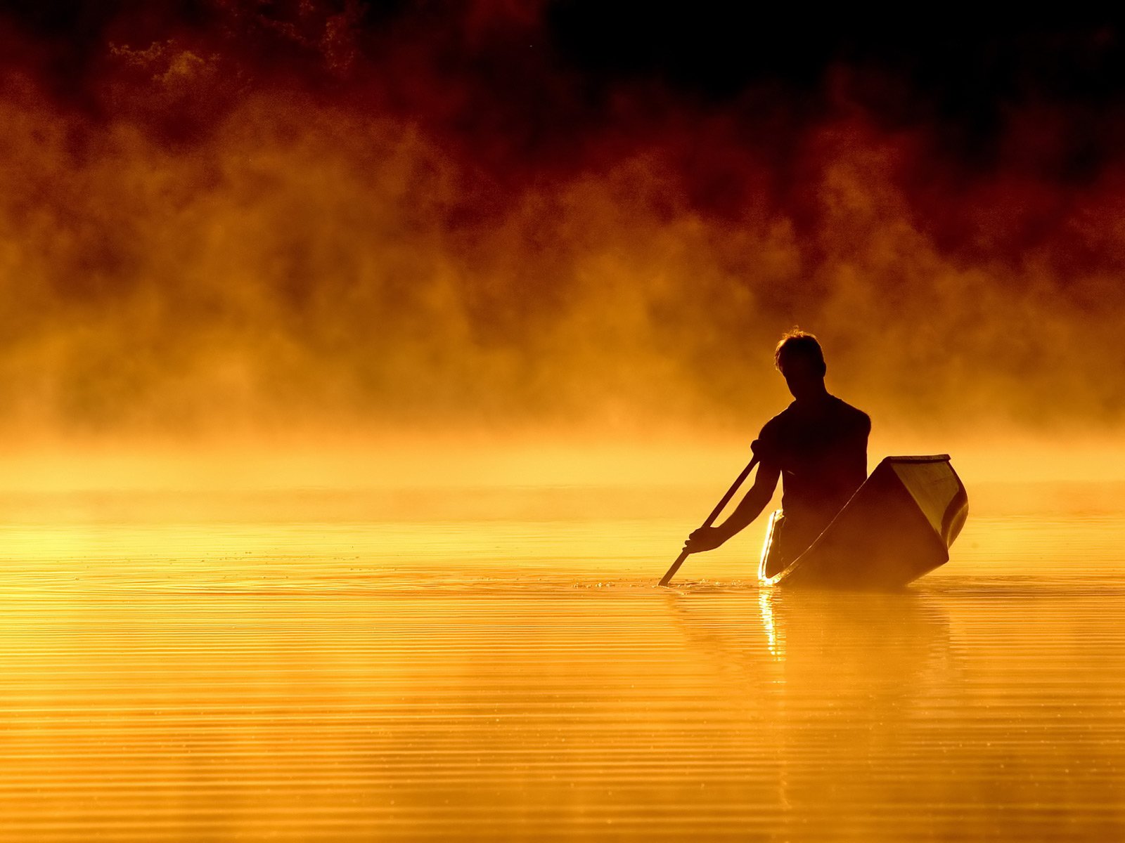 Photography of a person paddling a canoe on calm water, surrounded by golden mist at sunrise or sunset.