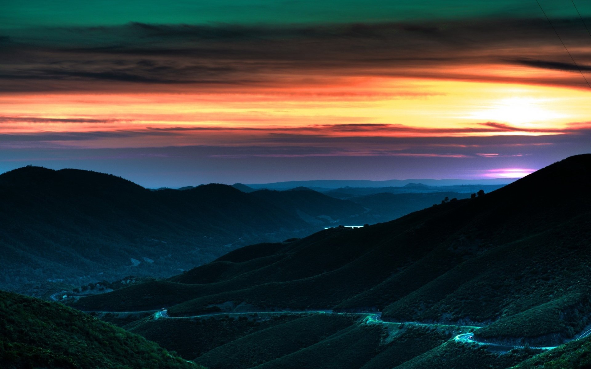 A vibrant sunset casts colorful light over rolling hills and winding roads in a serene nature landscape.