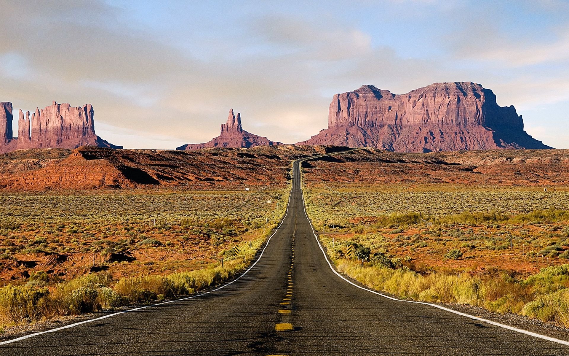 A long, straight road stretches through a desert landscape, flanked by majestic rock formations, showcasing the beauty of man-made infrastructure amidst natural scenery.
