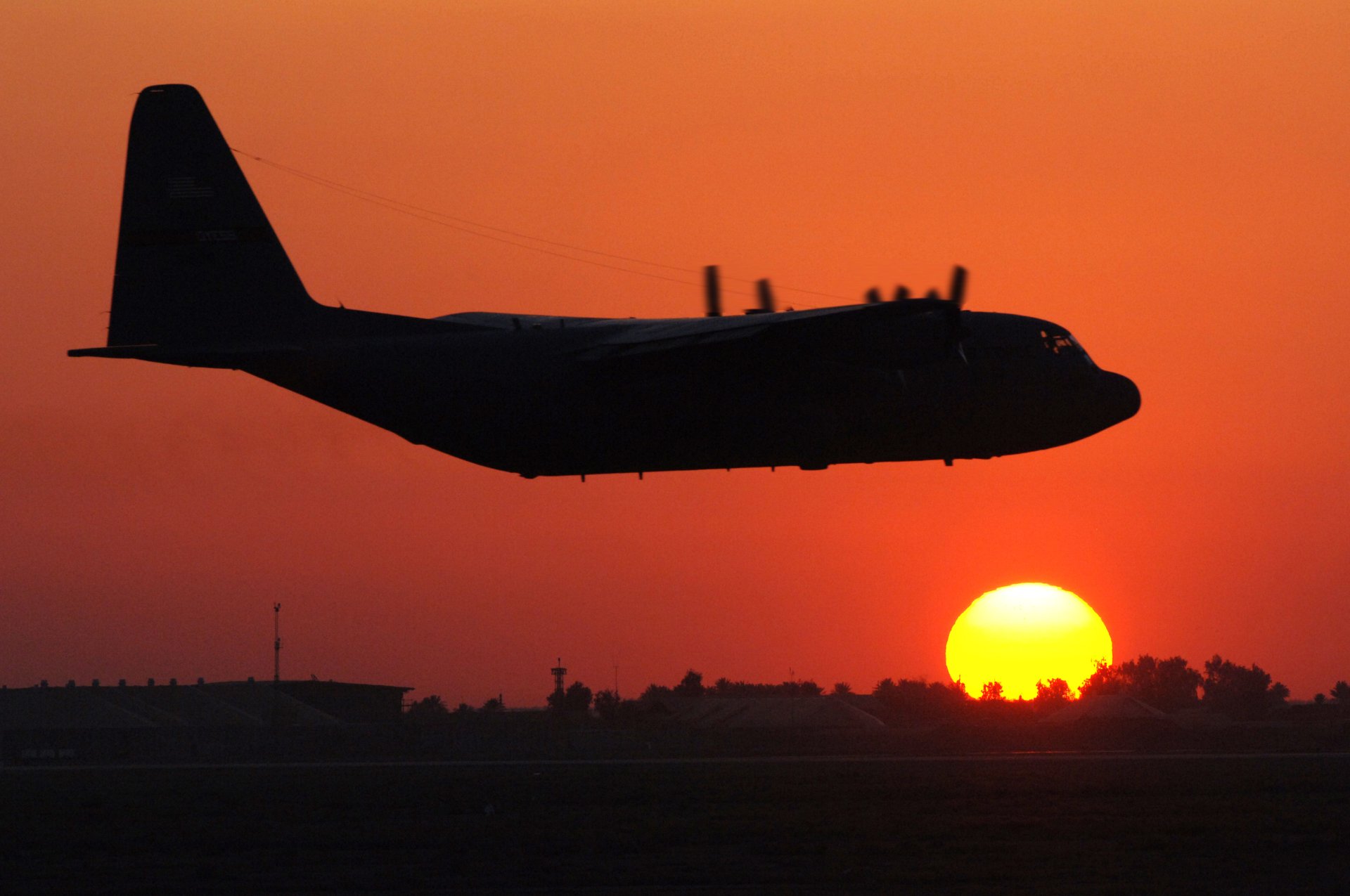 Silhouette of a military Lockheed C-130 Hercules flying across an orange sunset with the sun near the horizon.