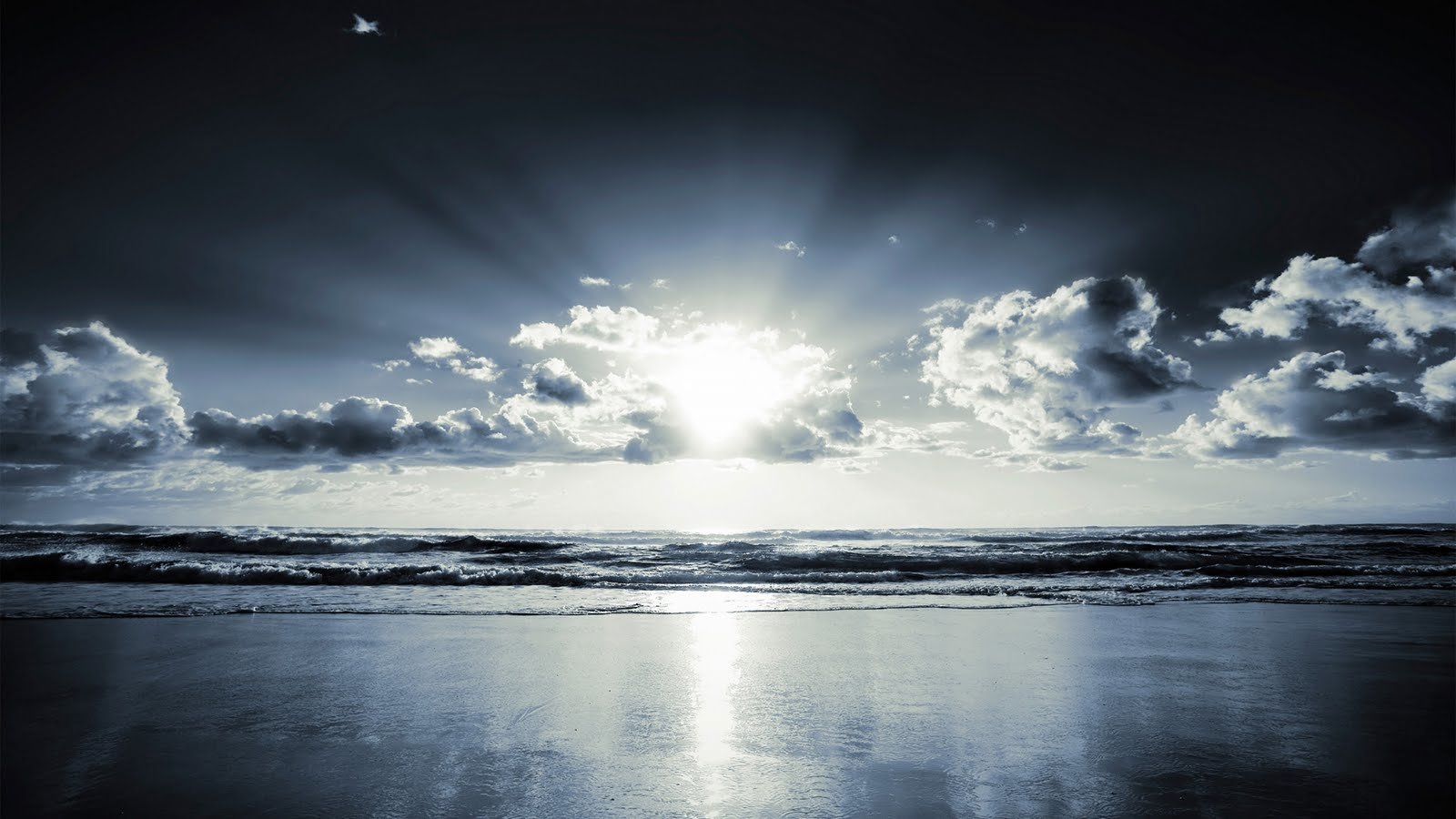 Scenic nature view of the sun shining through clouds over a calm ocean with its reflection on the wet sandy shore.