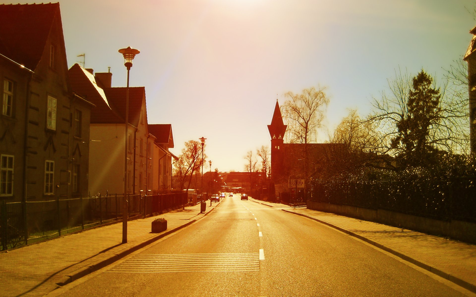 A sunlit road stretches through a residential area, lined with houses and trees, leading toward a clock tower in the distance, showcasing a peaceful urban scene.