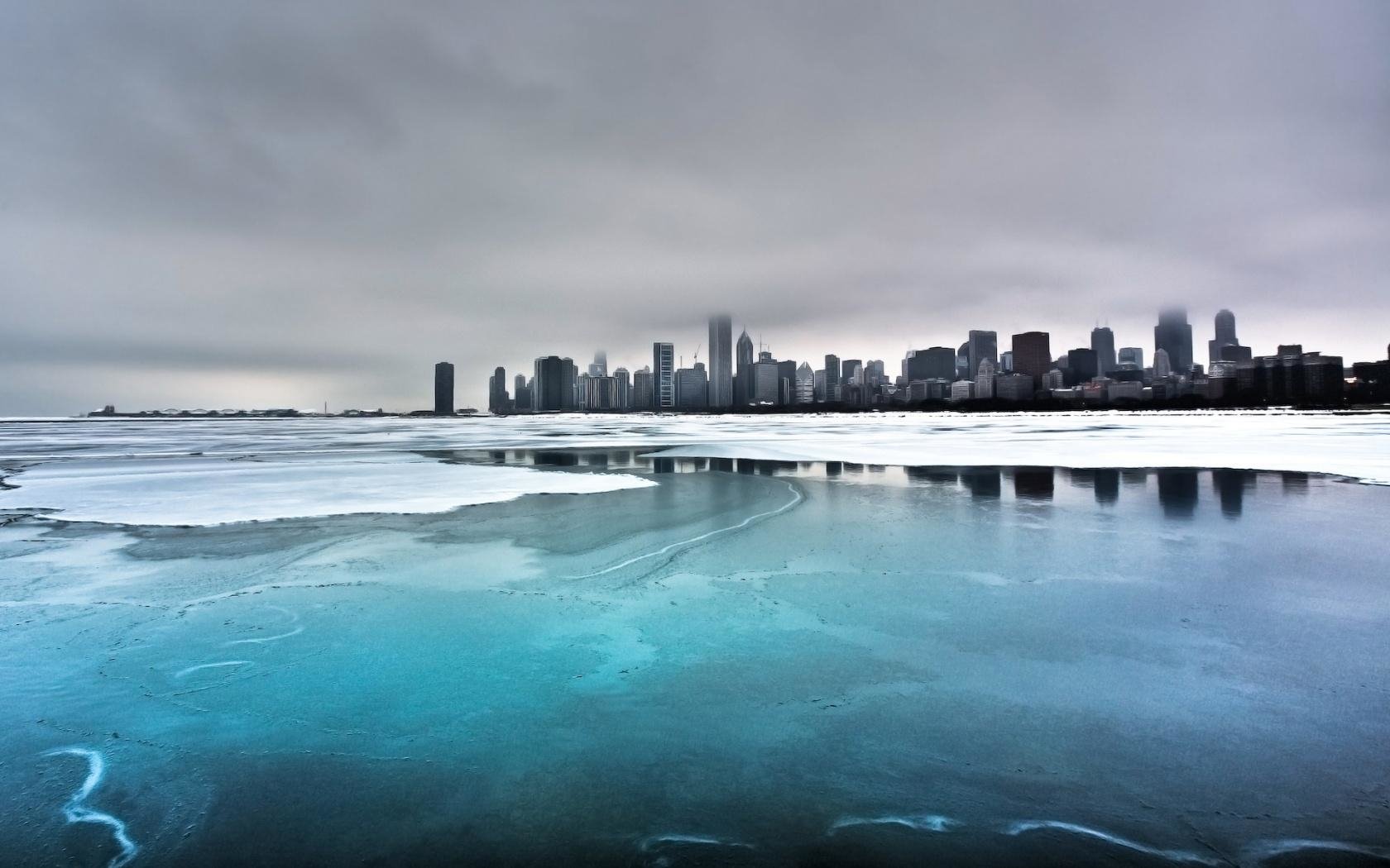 A city skyline under a cloudy sky with a large expanse of partially frozen water in the foreground, showcasing a man-made urban waterfront.