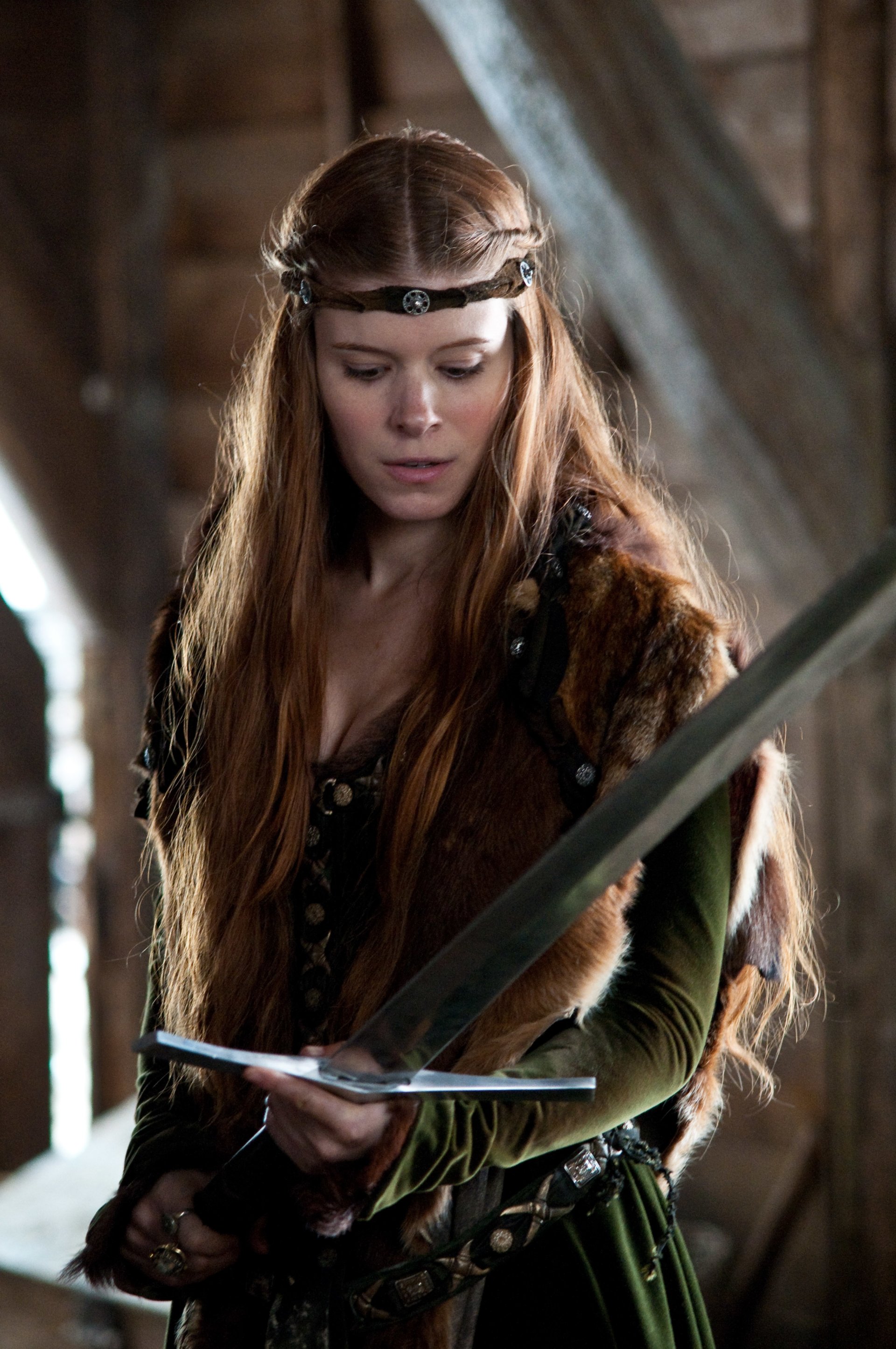 Still from the movie Ironclad: a red-haired medieval woman in a fur-trimmed gown studies a longsword inside a dim timber hall.
