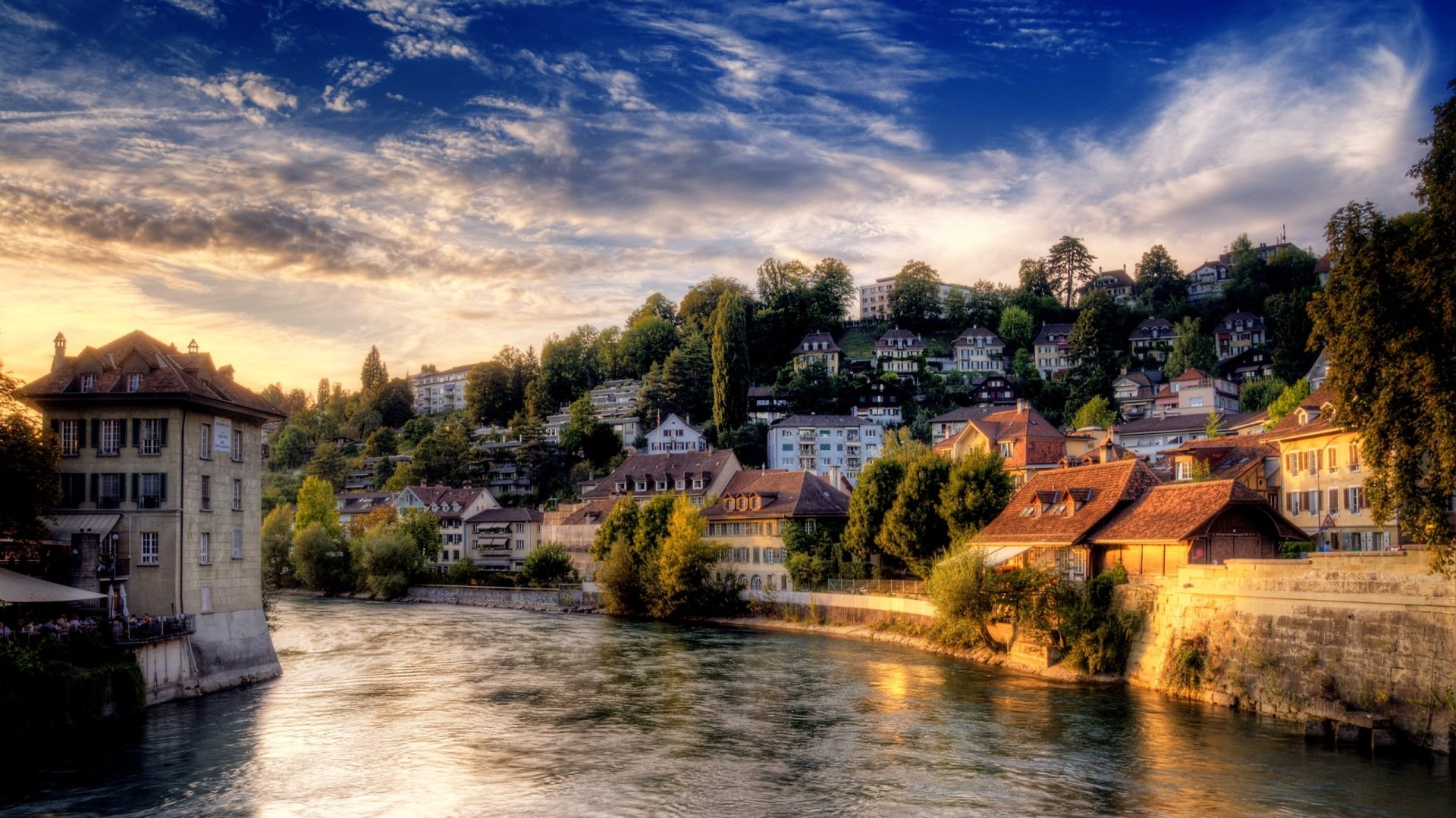 A stunning HDR photograph showcases a riverside view at sunset, highlighting charming houses nestled among lush greenery under a vibrant sky.