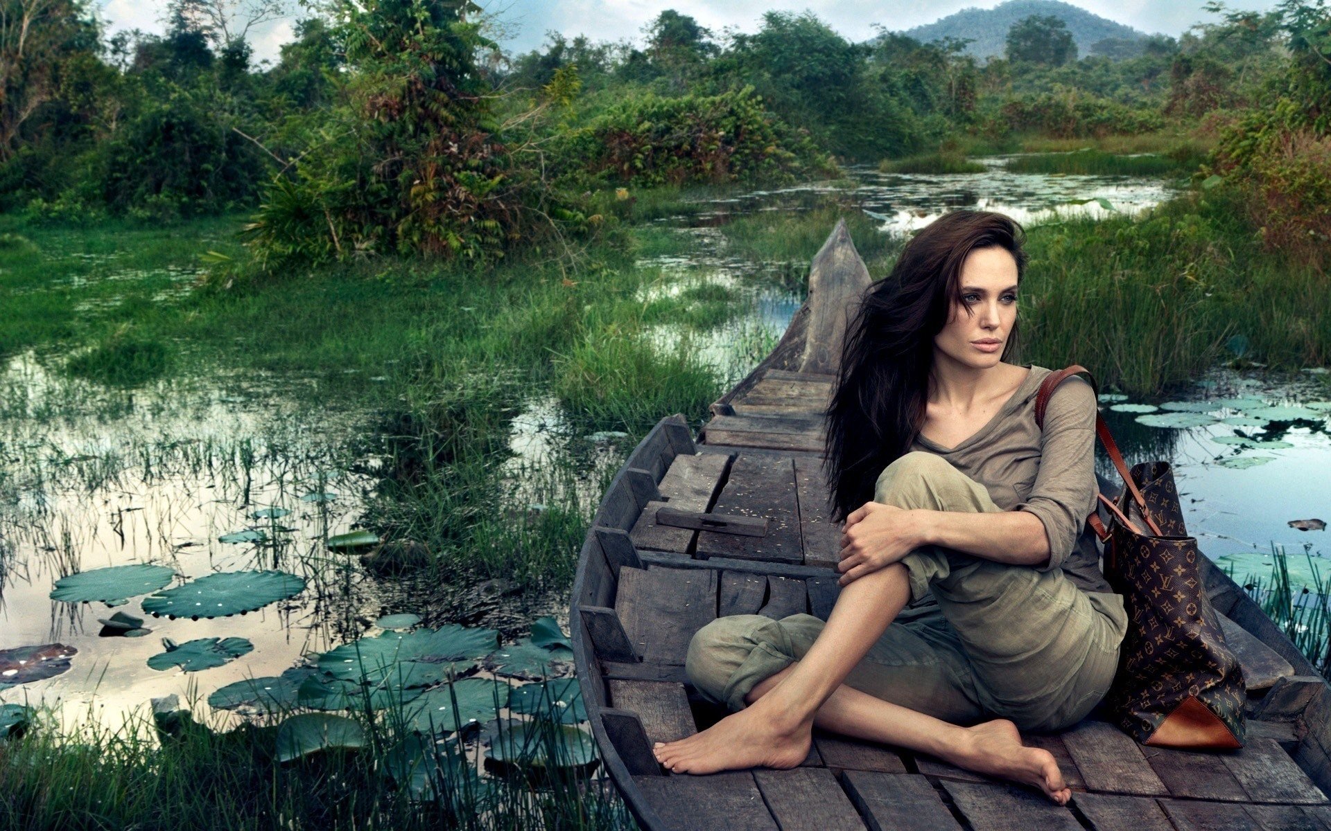 Angelina Jolie sits contemplatively on a wooden boat amidst a lush, green landscape, surrounded by tranquil water and distant mountains.