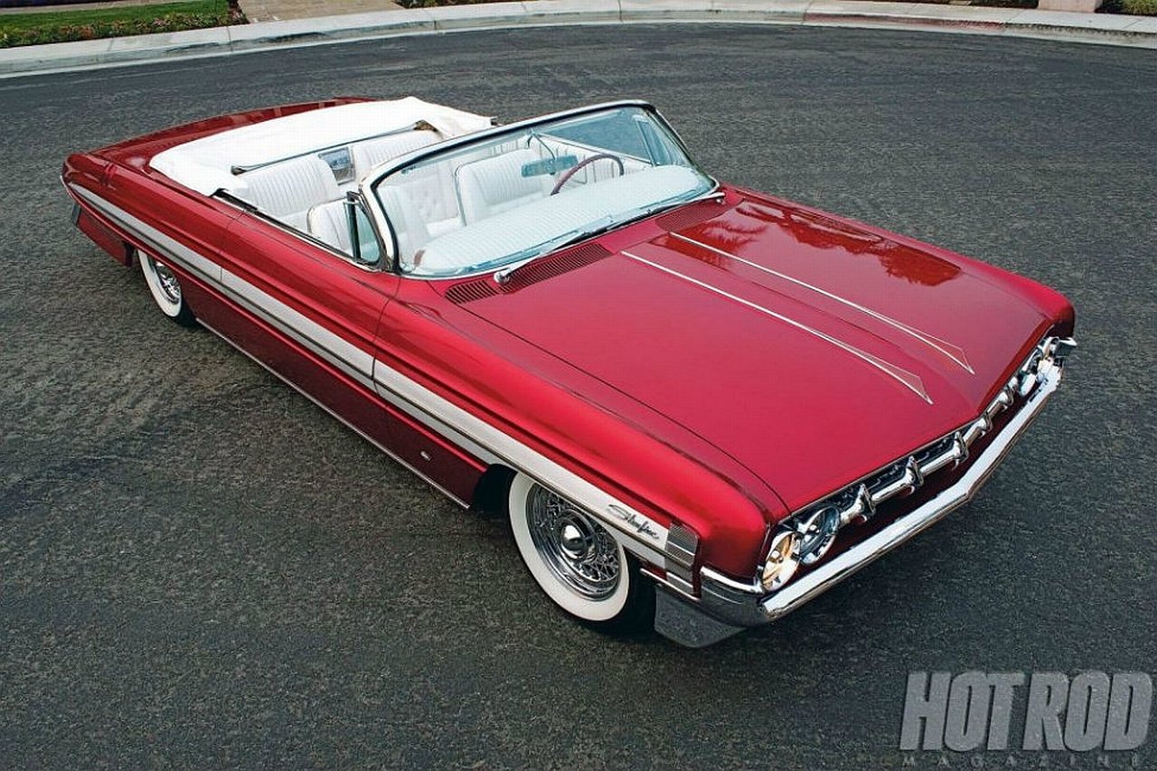 A shiny red vintage Oldsmobile convertible with white interior parked on asphalt.