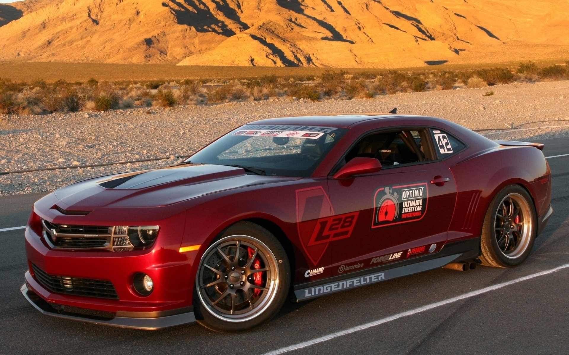 Red Chevrolet Camaro vehicle with racing decals and aftermarket wheels parked on a desert road at sunset.