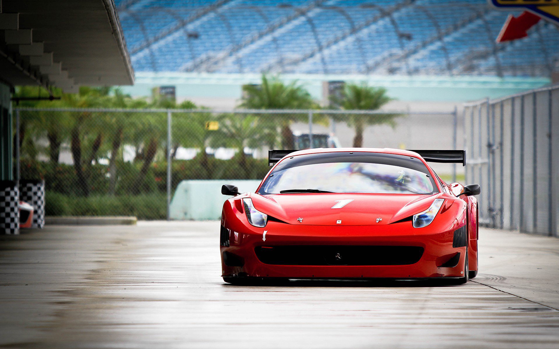 A red Ferrari vehicle is parked on a paved area with a racetrack and palm trees visible in the background.
