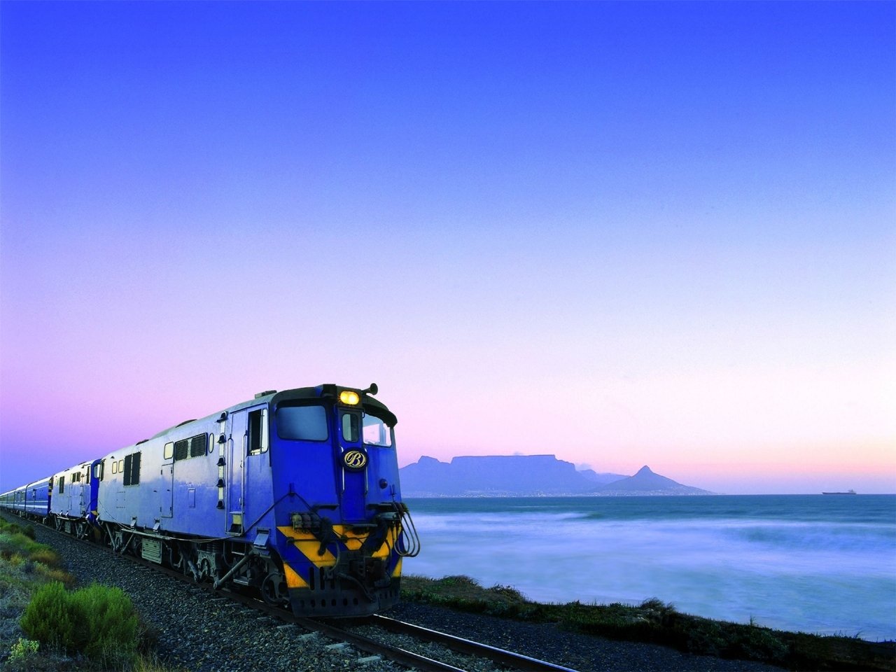 A blue train travels along the tracks by the coast, with a stunning sunset backdrop featuring a mountain silhouette and a tranquil sea.