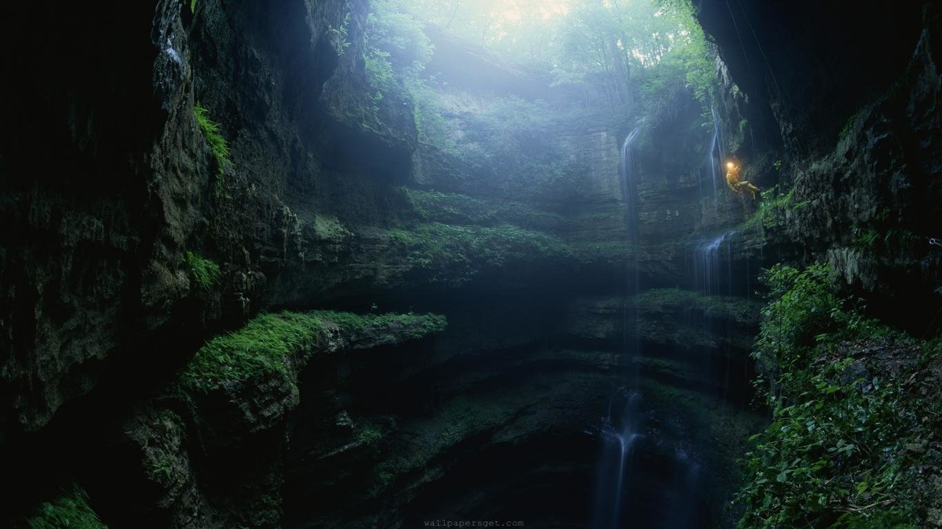 A lush, green cave opening reveals sunlight streaming down into the depths, highlighting natural rock formations and vegetation.