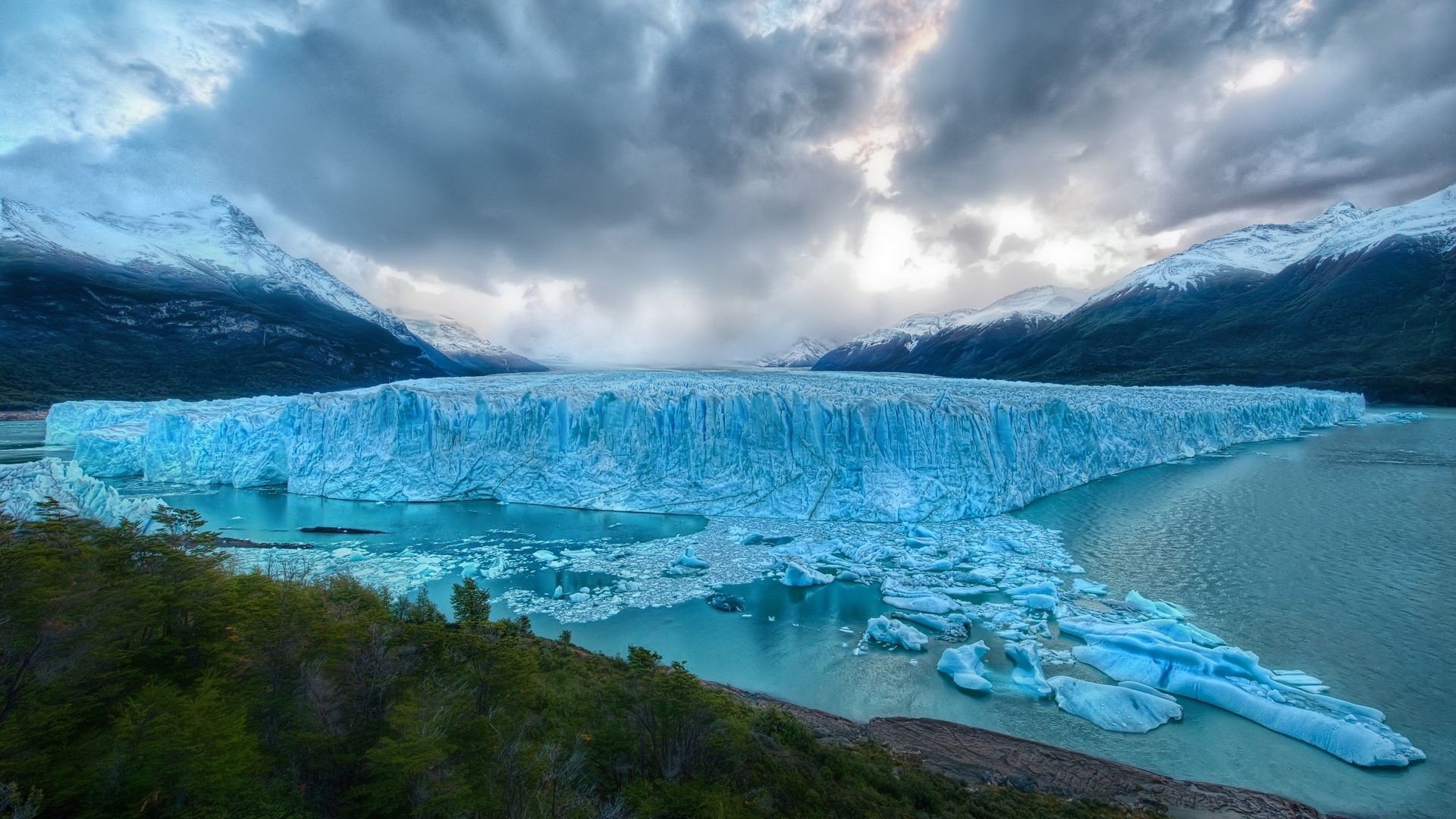 HDR nature view of a massive blue glacier meeting an icy lake, chunks of ice floating beneath dramatic cloudy skies and a forested shore.