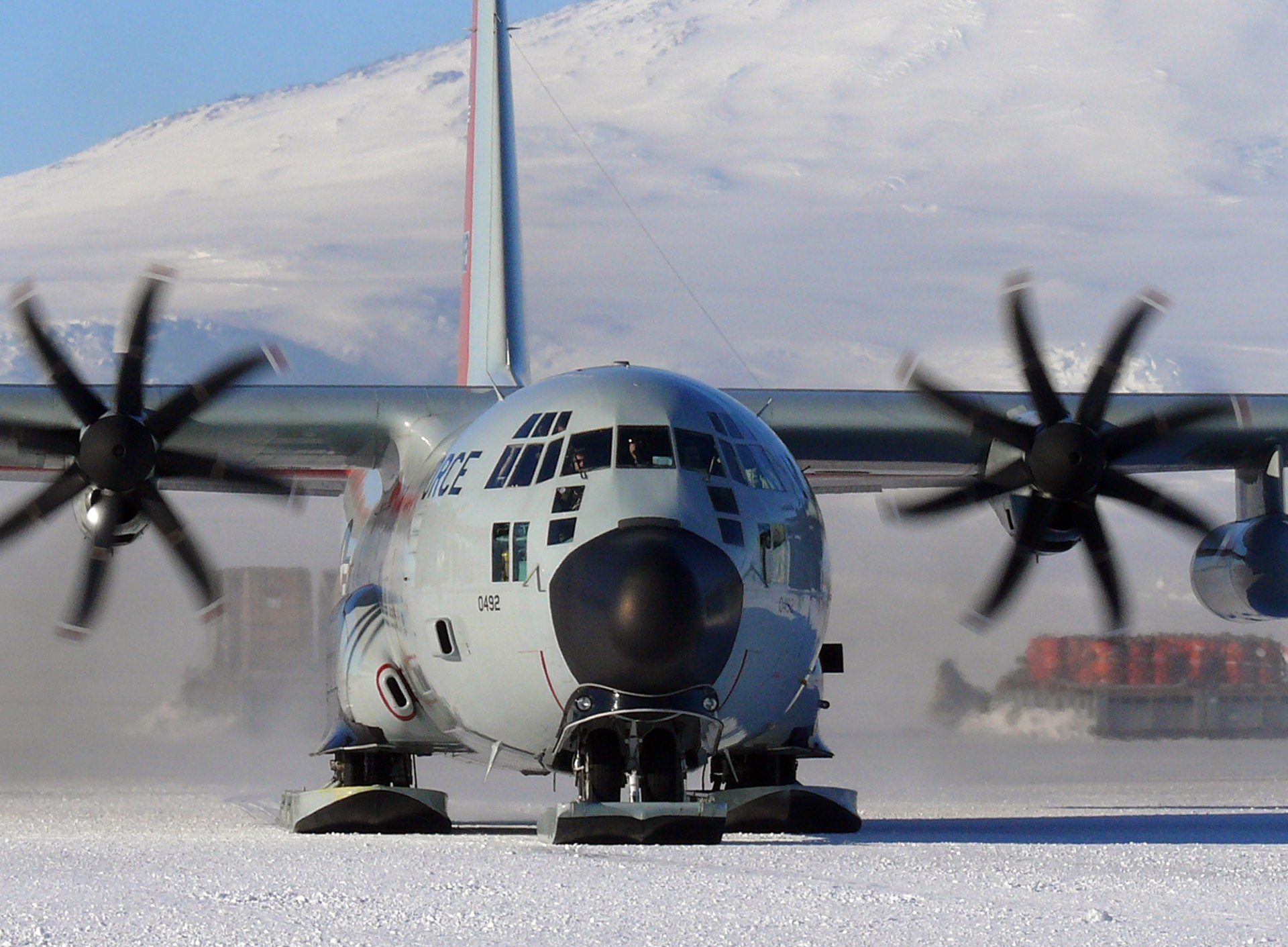 Military C-130 Hercules aircraft equipped with skis for snow-covered terrain, taxiing on a snowy surface with its propellers spinning.