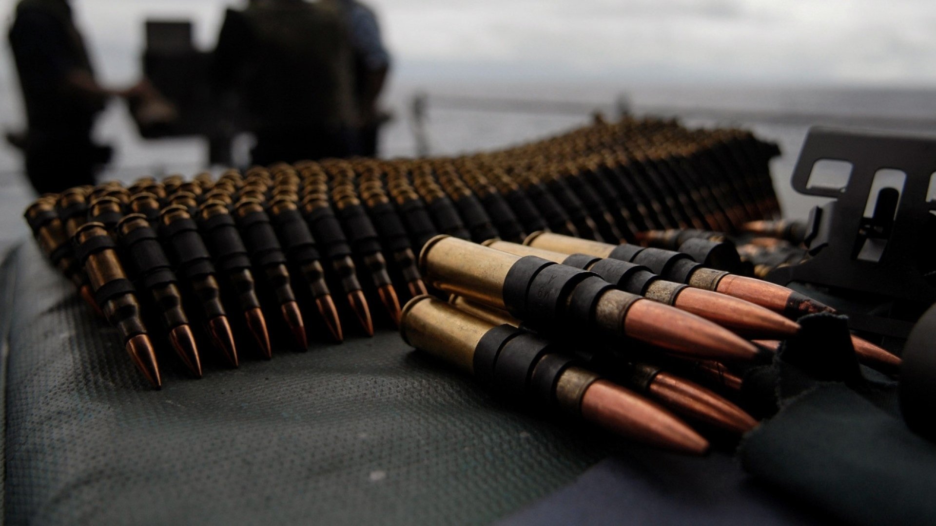 Close-up of man-made linked rifle bullets and brass casings on a surface, with blurred figures and a distant horizon in the background.