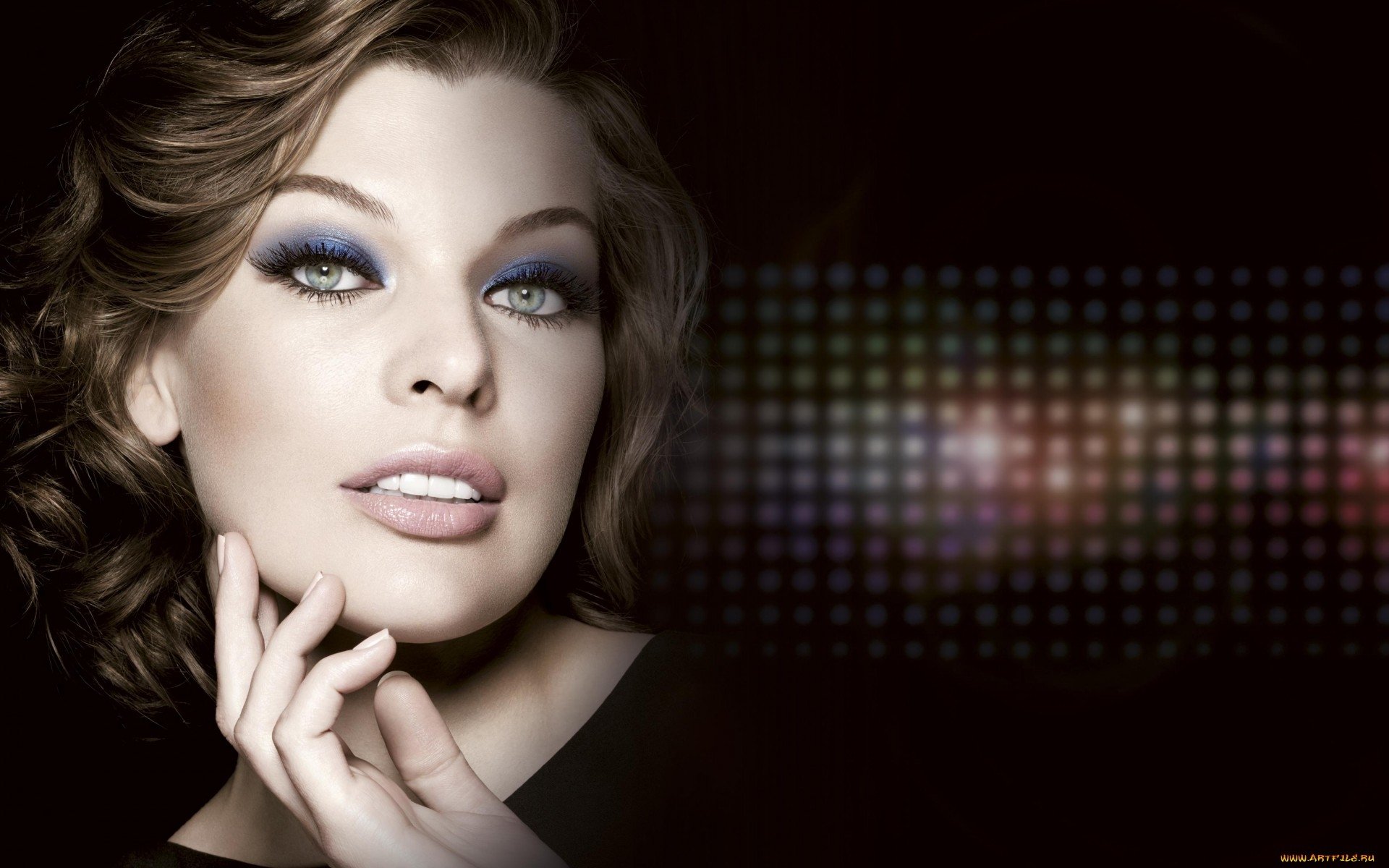 Close-up portrait of celebrity Milla Jovovich with smoky eye makeup and softly curled hair against a dark, abstract dotted background.
