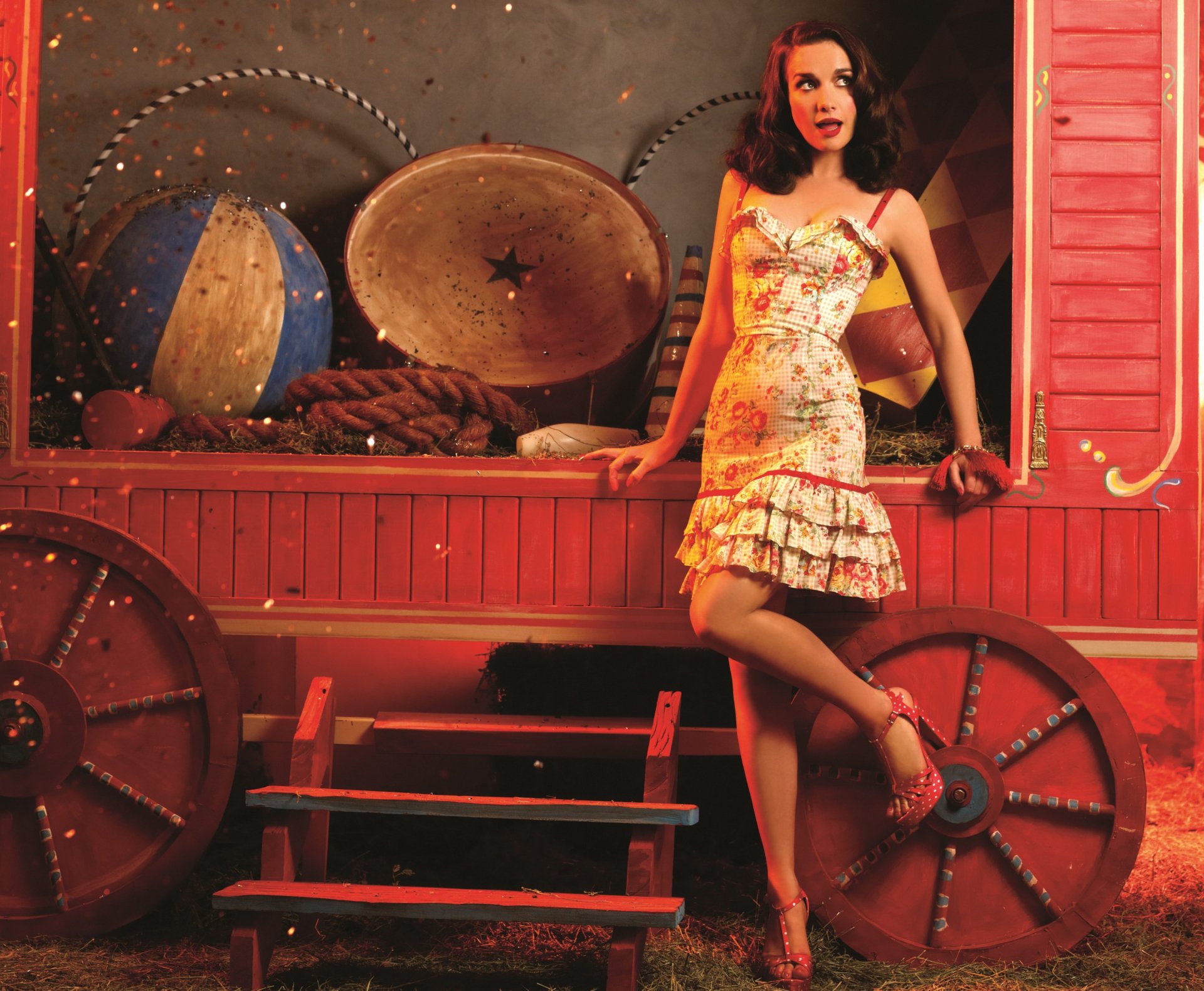 Natalia Oreiro poses in a floral dress on a vintage wagon set, surrounded by musical instruments, evoking a warm, artistic atmosphere.