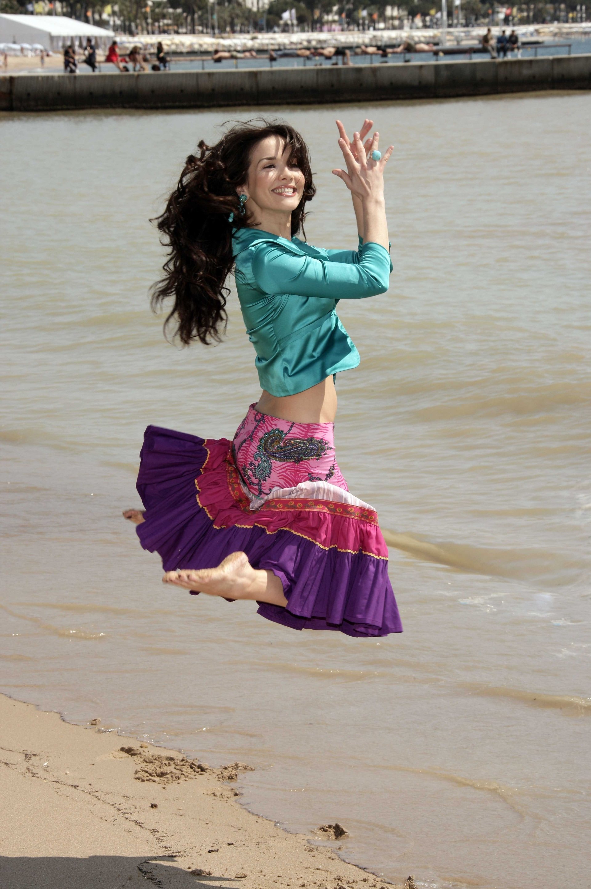 Natalia Oreiro joyfully leaps on a beach, wearing a colorful skirt and teal blouse, expressing music and dance by the water.