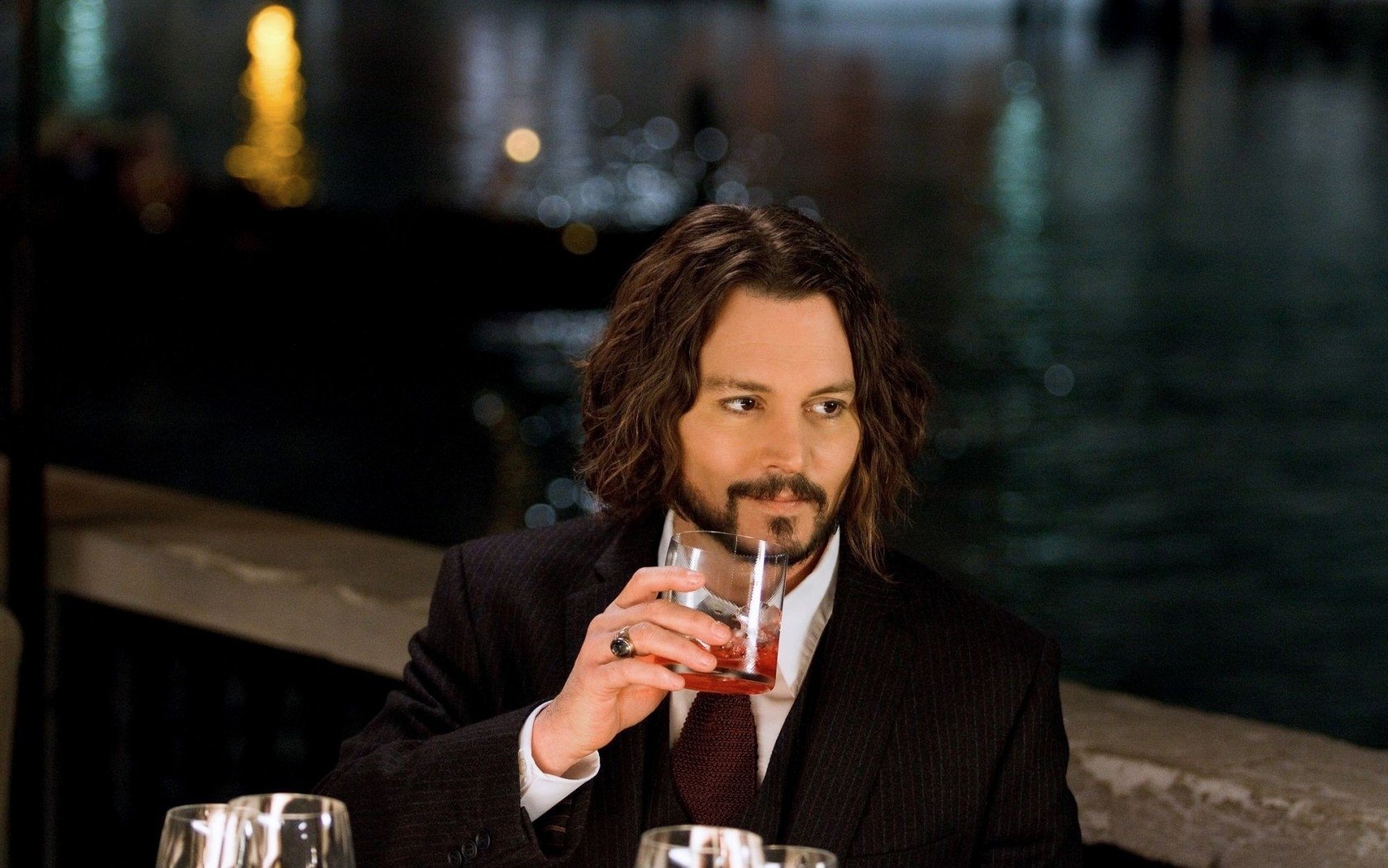 Johnny Depp sits at a table by the water, dressed in a sleek suit, casually sipping a drink with an intriguing expression. The background features softly lit reflections on the water.