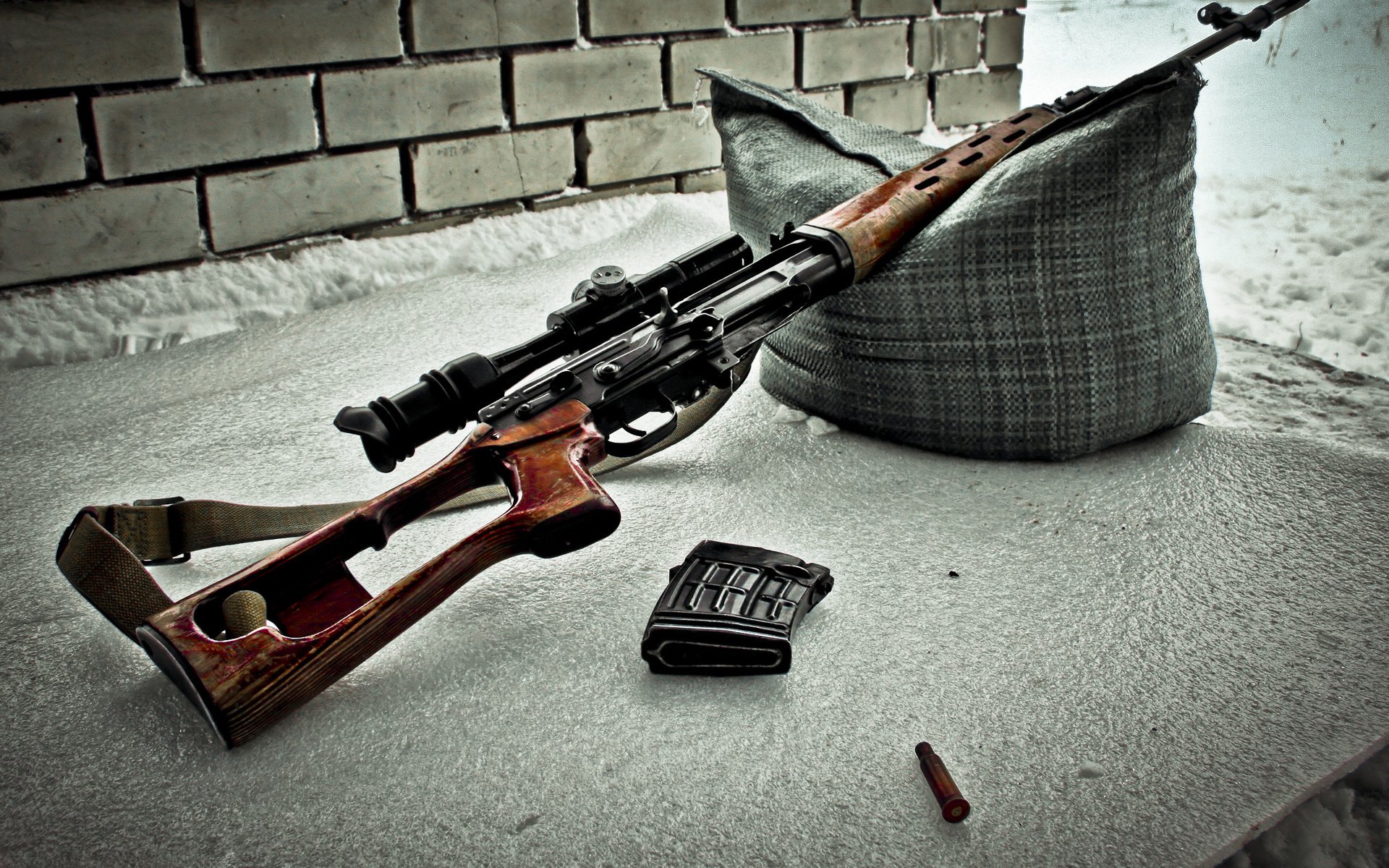 A sniper rifle rests on a surface with a cushion, accompanied by a magazine and a bullet, set against a backdrop of bricks, highlighting a military scene.